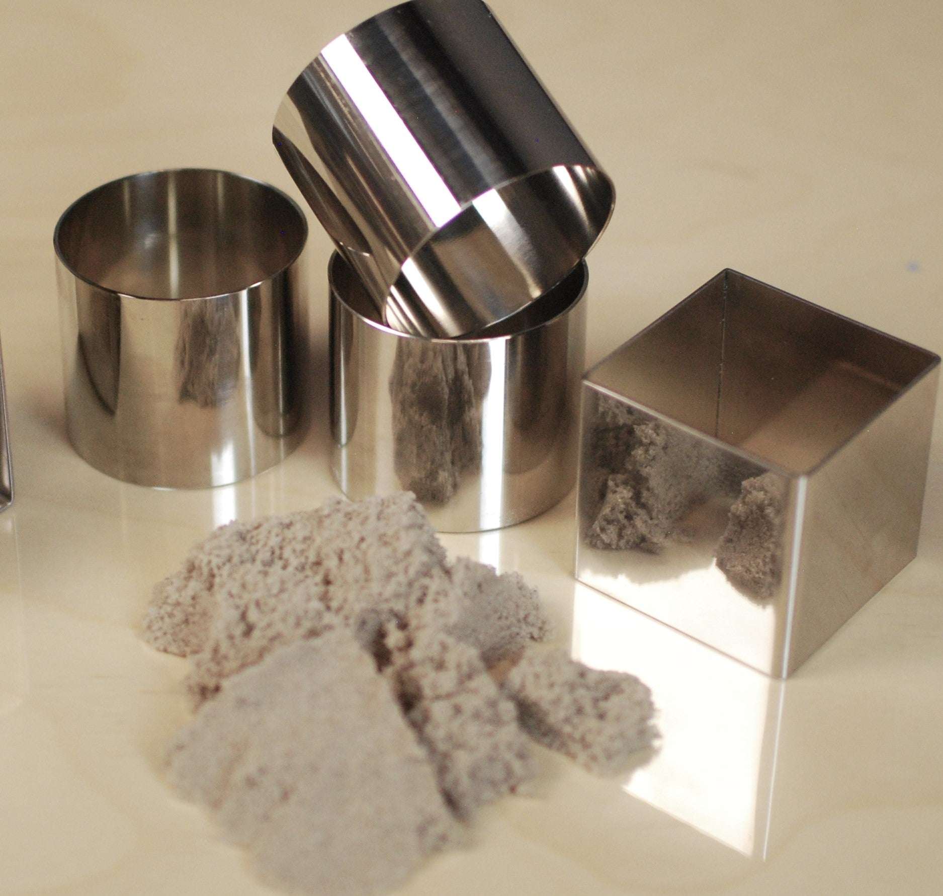 Set of six durable stainless steel square and cylindrical molds with kinetic sand on a light surface.