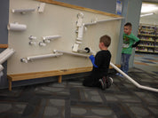 Children playing with white magnetic ramps and tubes on a large, wall-mounted wooden board in a library.