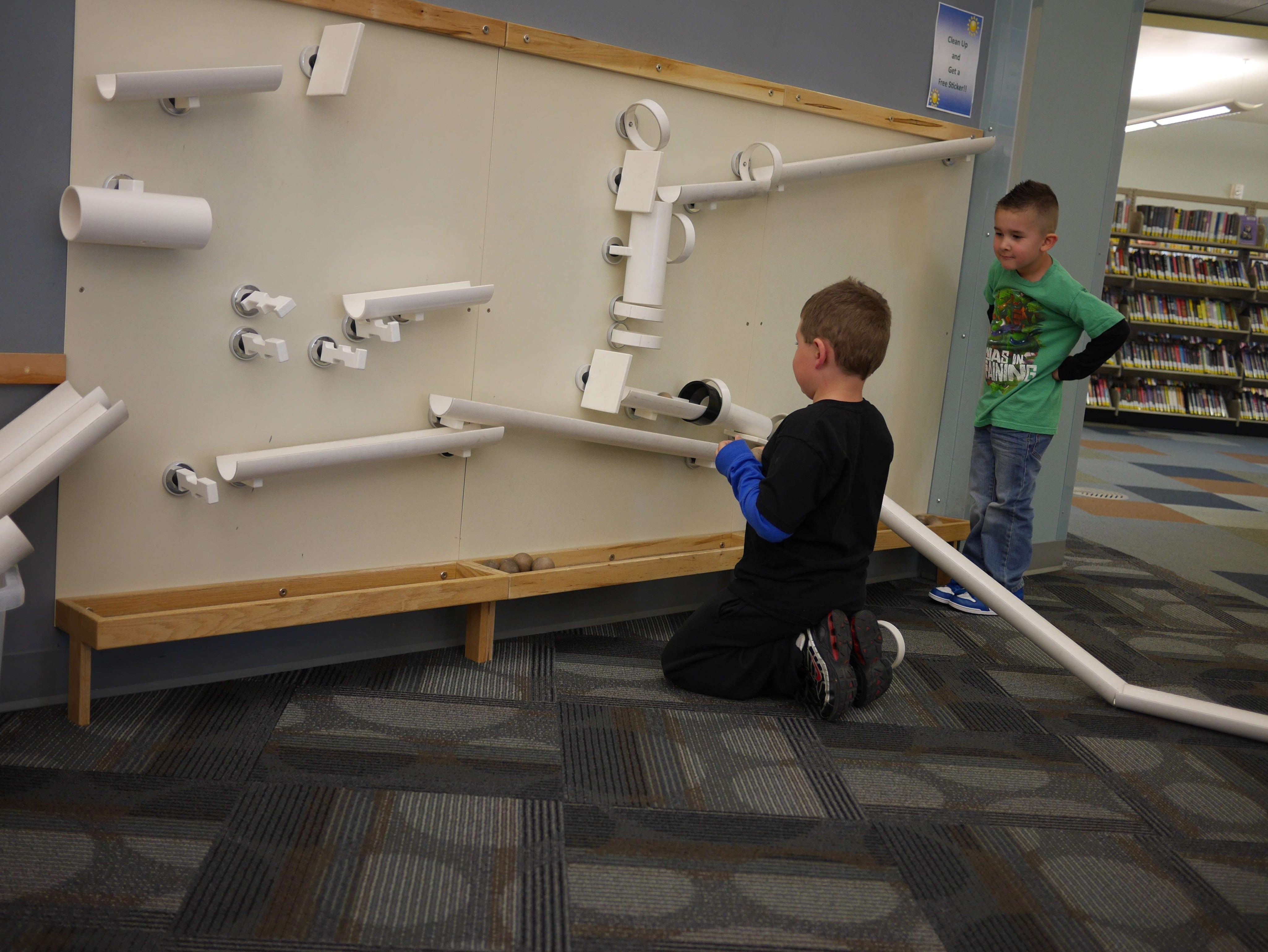 Children playing with white magnetic ramps and tubes on a large, wall-mounted wooden board in a library.
