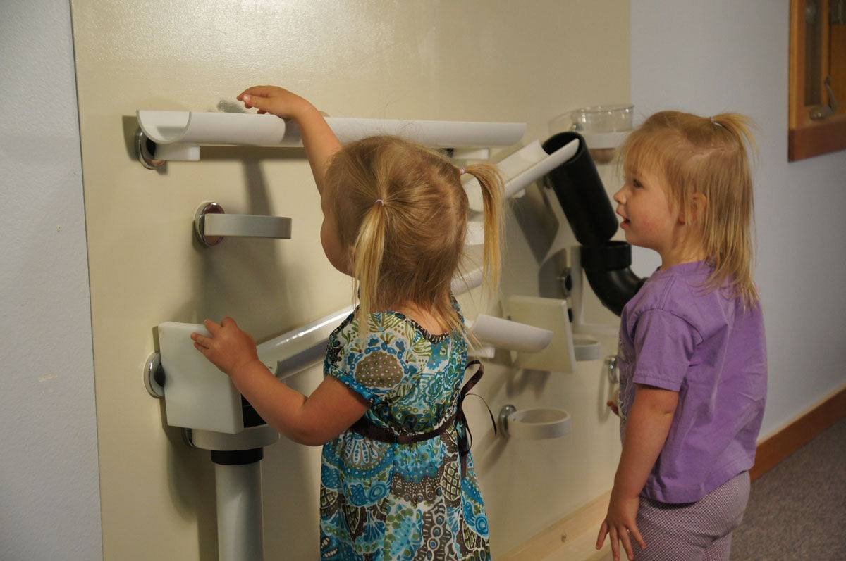 Children playing with white magnetic ramps and tracks on a beige vertical museum wall.