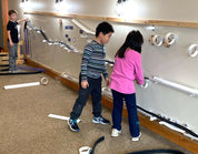 Children playing with magnetic ramps and tracks on a large, light-colored vertical wall in a classroom setting.