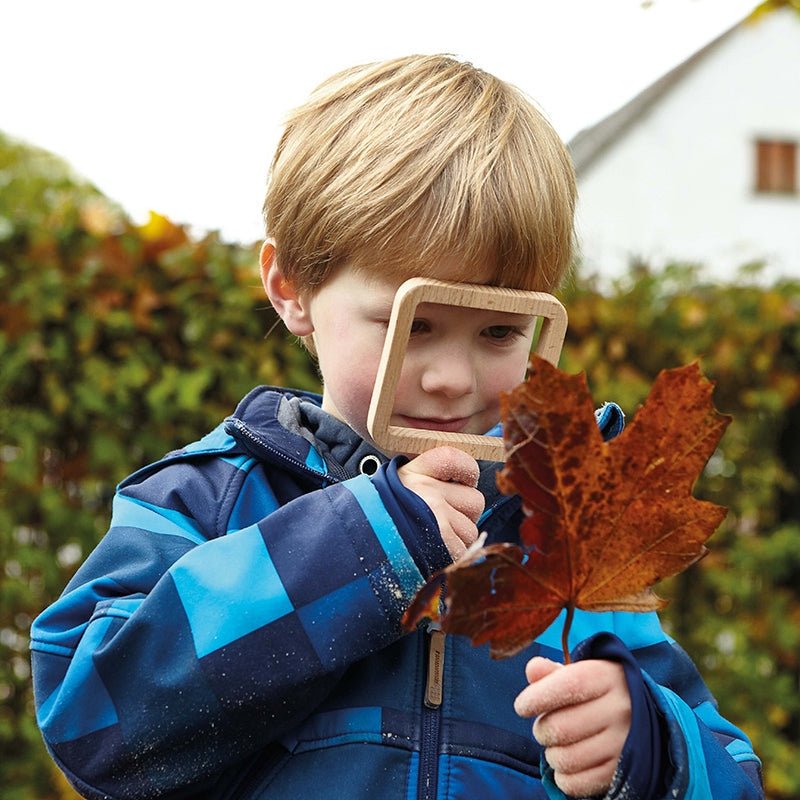 Child looking through a square beech wood viewer at a brown autumn leaf outdoors.