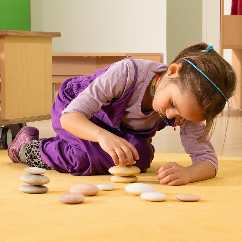 A young girl stacking smooth, natural-colored sorting stones on a yellow rug for educational play.