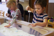 Child pouring white sand between two small, rustic coconut shell spoons in a classroom sensory table.