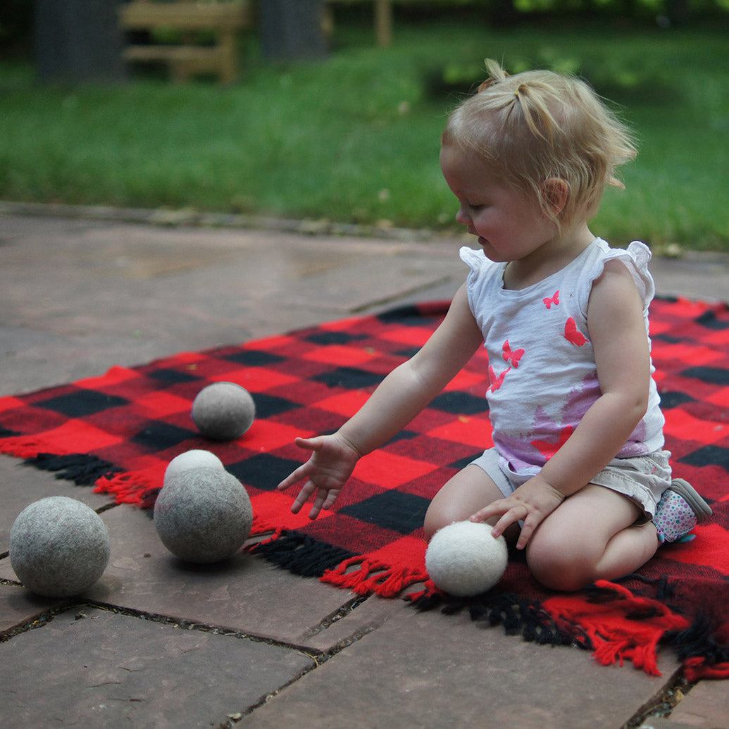Toddler playing with soft, undyed natural wool balls on a red and black plaid blanket outdoors.