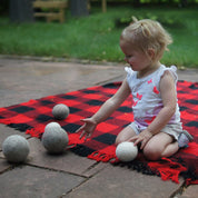 Toddler playing with soft, undyed natural wool balls on a red and black plaid blanket outdoors.