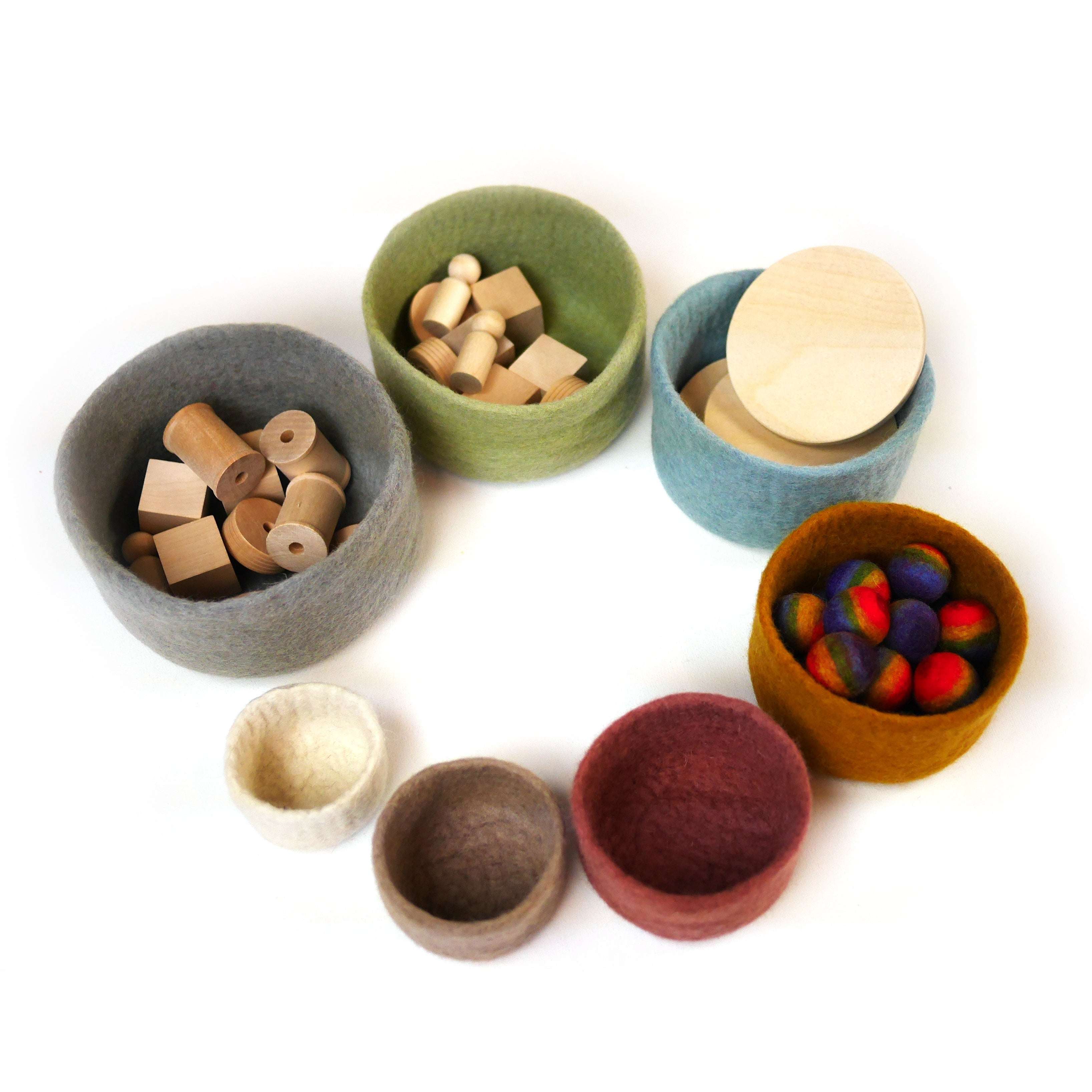 Set of seven nested wool bowls in earthy tones, filled with wooden loose parts and colorful felt balls on a white background.