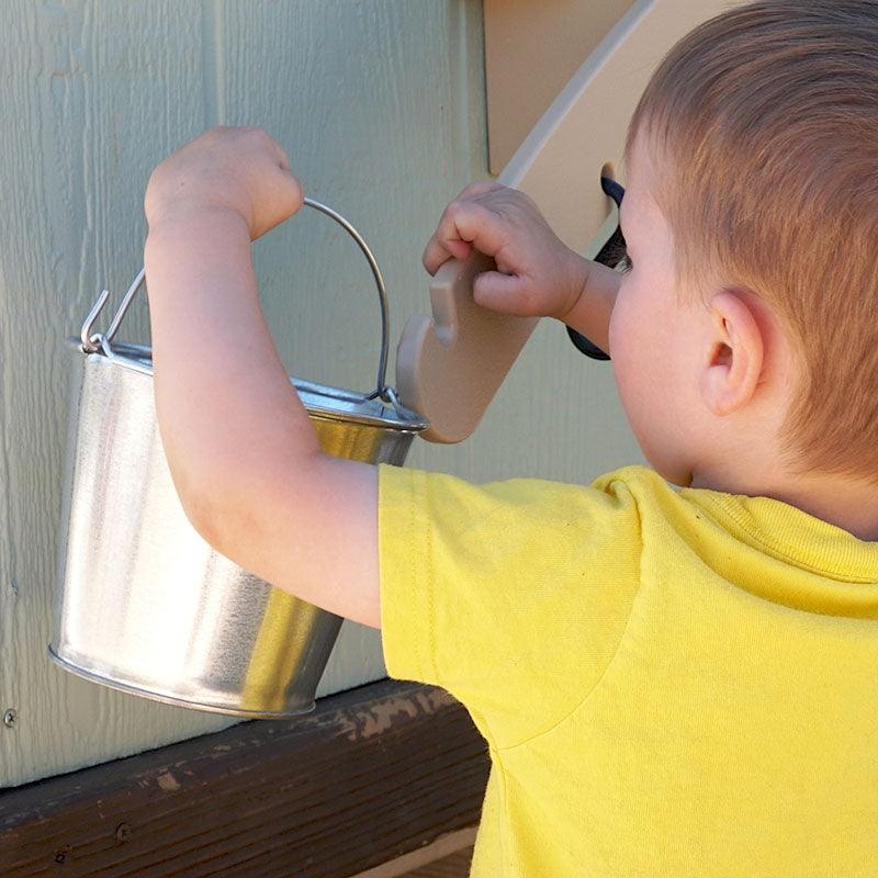 Child in a yellow shirt hanging a metal pail on a beige outdoor balance scale mounted to a wall.