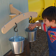 Child playing with a wall-mounted outdoor balance scale featuring two metal pails hanging from black carabiners.