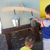 Tan wooden balance scale mounted on a wall with two hanging metal pails for children to explore weight and mass.