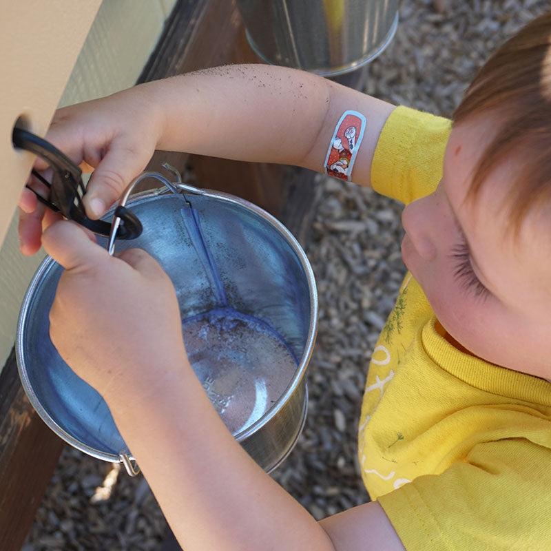 Child in a yellow shirt attaching a metal pail to a wall-mounted outdoor balance scale using a black carabiner.