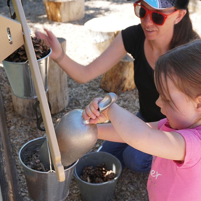 Child using a metal scoop to fill a hanging metal bucket on a wooden outdoor balance scale with pine cones.