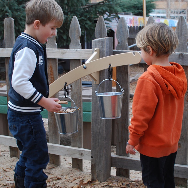 Two children playing with a wooden balance scale mounted to a fence, featuring two metal pails hanging from carabiners.