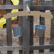 Tan outdoor balance scale mounted on a wooden fence with two hanging metal pails attached by black carabiners.