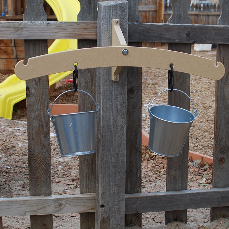 Tan outdoor balance scale mounted on a wooden fence with two hanging metal pails attached by black carabiners.