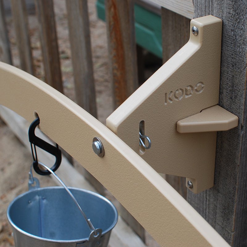 Tan plastic outdoor balance scale mounted to a wooden post with a metal pail hanging from a black carabiner.