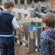 Children playing with a tan wooden balance scale mounted to a fence, featuring two metal pails hanging from carabiners.