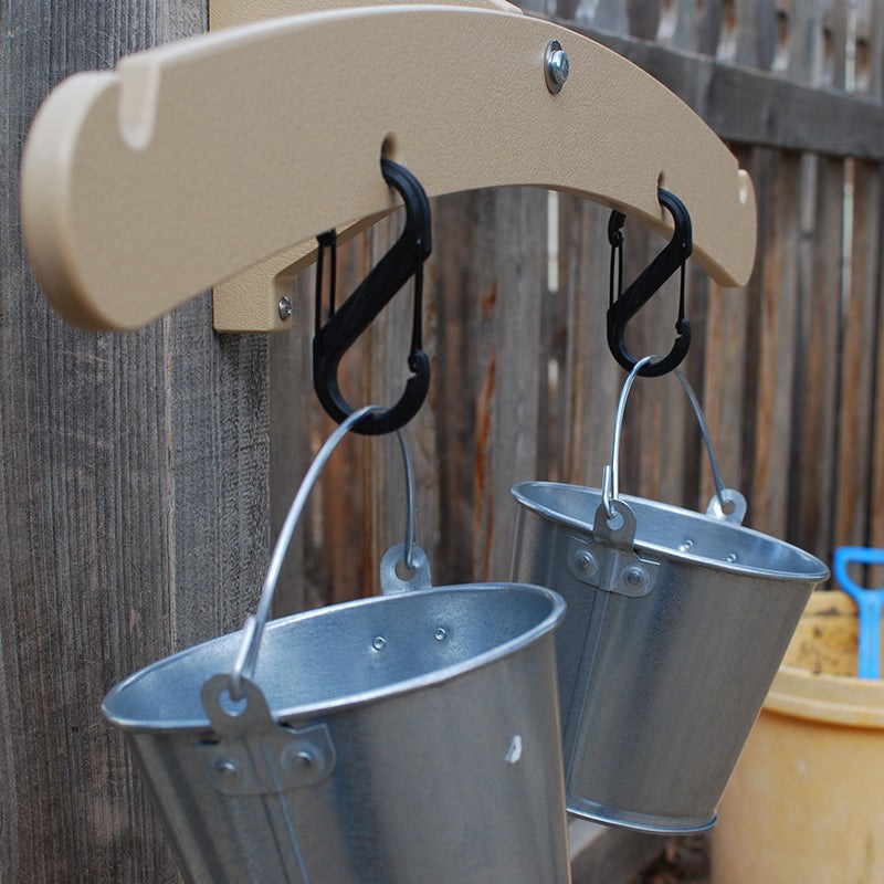 Beige outdoor balance scale mounted on a wooden fence with two hanging metal pails attached by black carabiners.