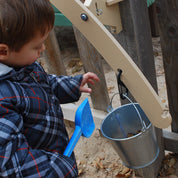Child playing with a beige outdoor balance scale mounted to a wooden fence, featuring a hanging metal pail and blue shovel.