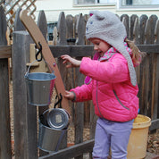 Child playing with a tan wooden outdoor balance scale mounted to a fence, featuring two hanging metal pails and carabiners.
