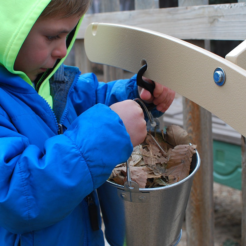 Child hanging a metal pail filled with leaves onto a beige, wall-mounted outdoor balance scale.