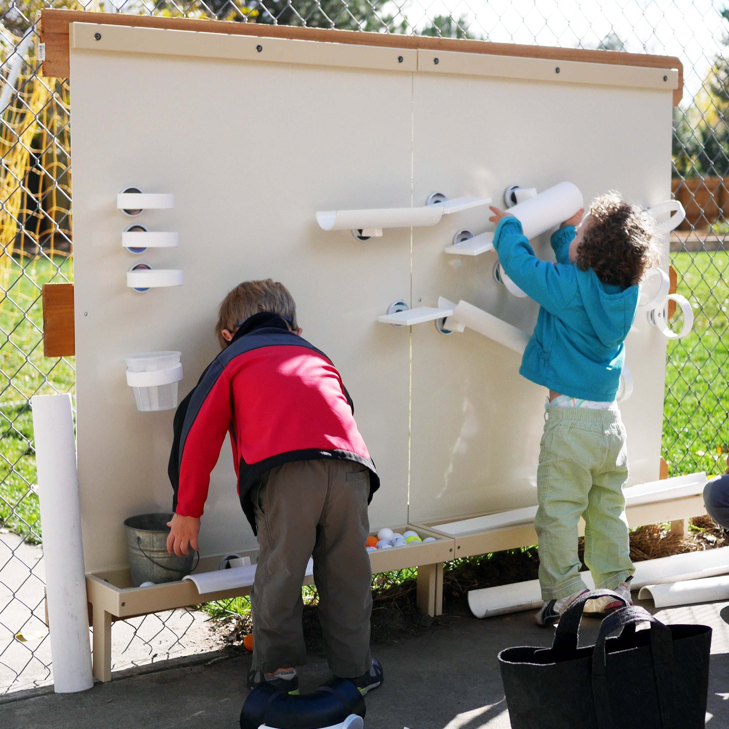 Children playing with a large, beige magnetic wall featuring white ramps, cups, and trays on a playground.