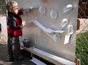 Child playing with a white outdoor magnetic wall featuring ramps, rings, and a metal tray for interactive STEM learning.