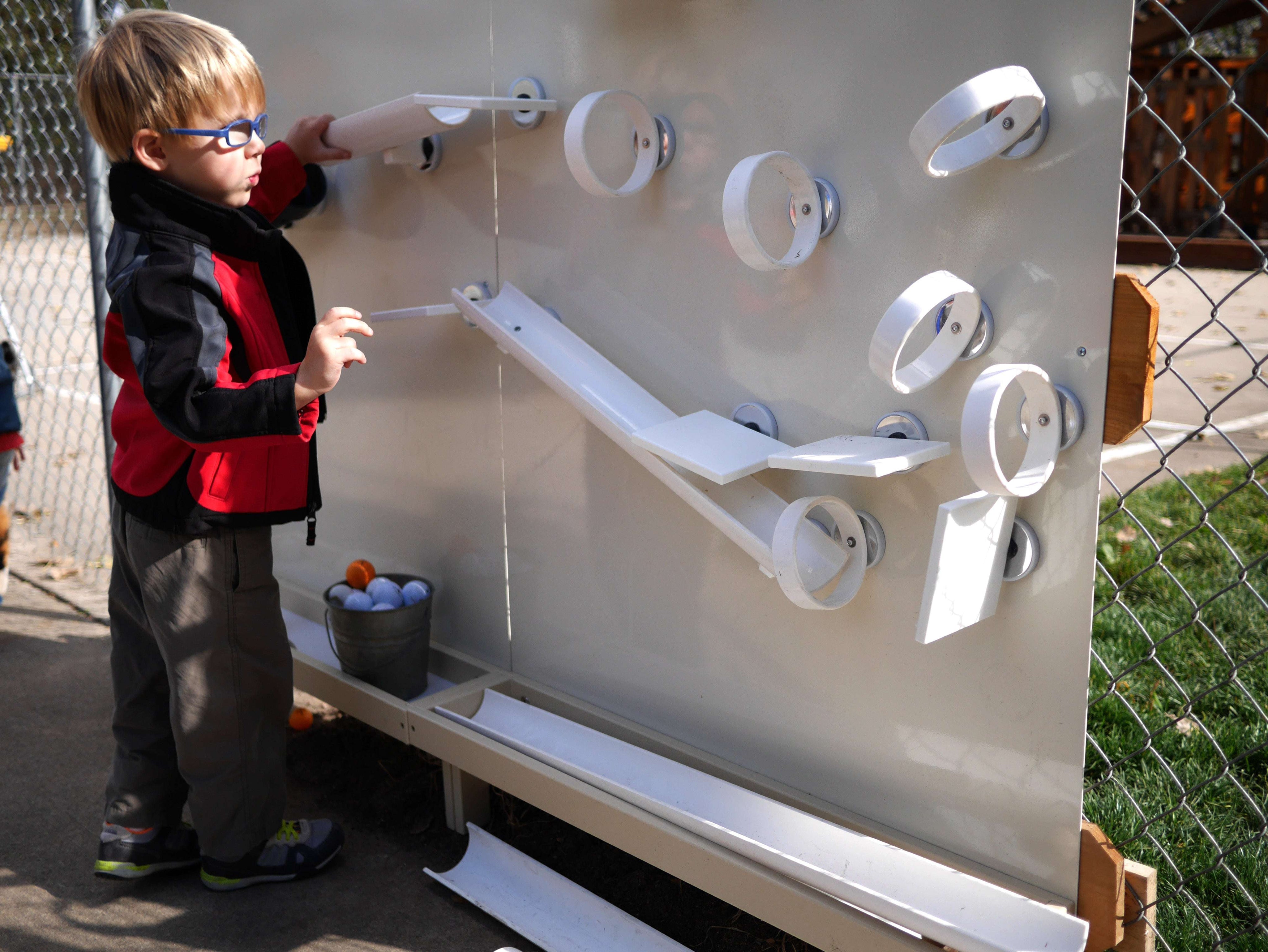 Child playing with a white outdoor magnetic wall featuring ramps, rings, and a metal tray for interactive STEM learning.