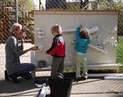 Children and teacher interacting with a large, beige metal outdoor magnet wall featuring white ramps and cups.
