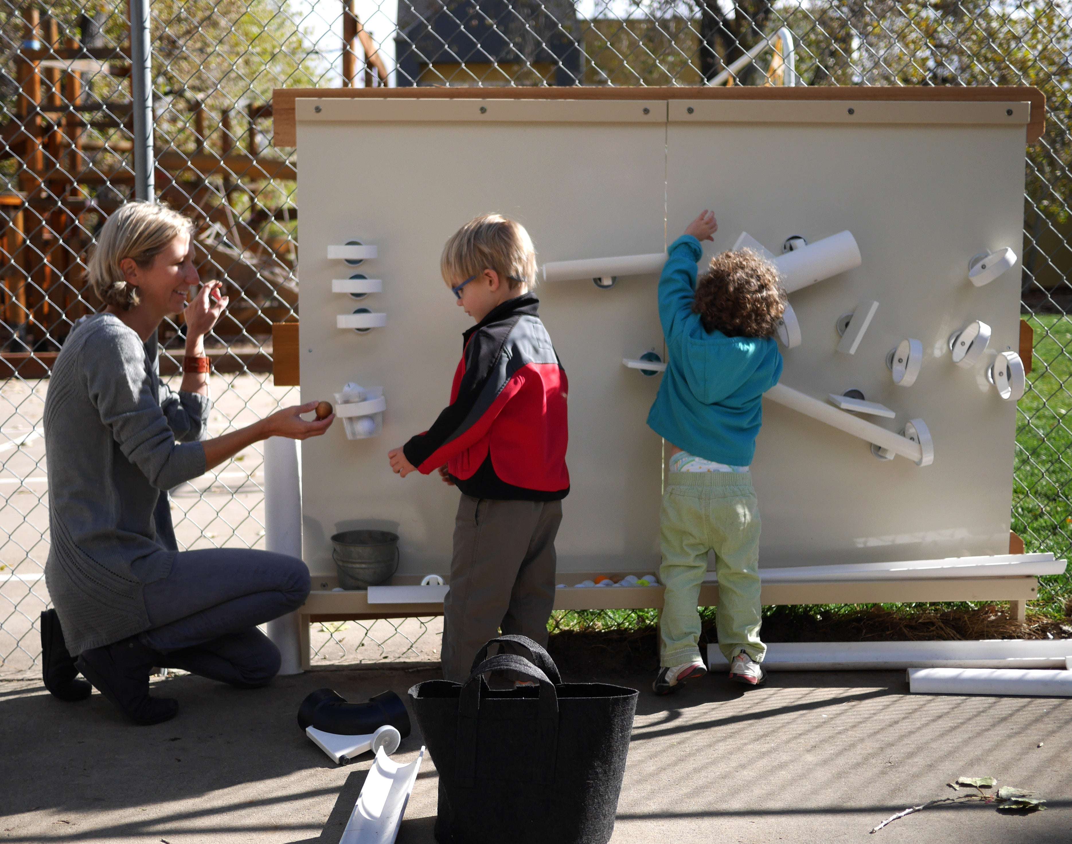 Children and teacher interacting with a large, beige metal outdoor magnet wall featuring white ramps and cups.