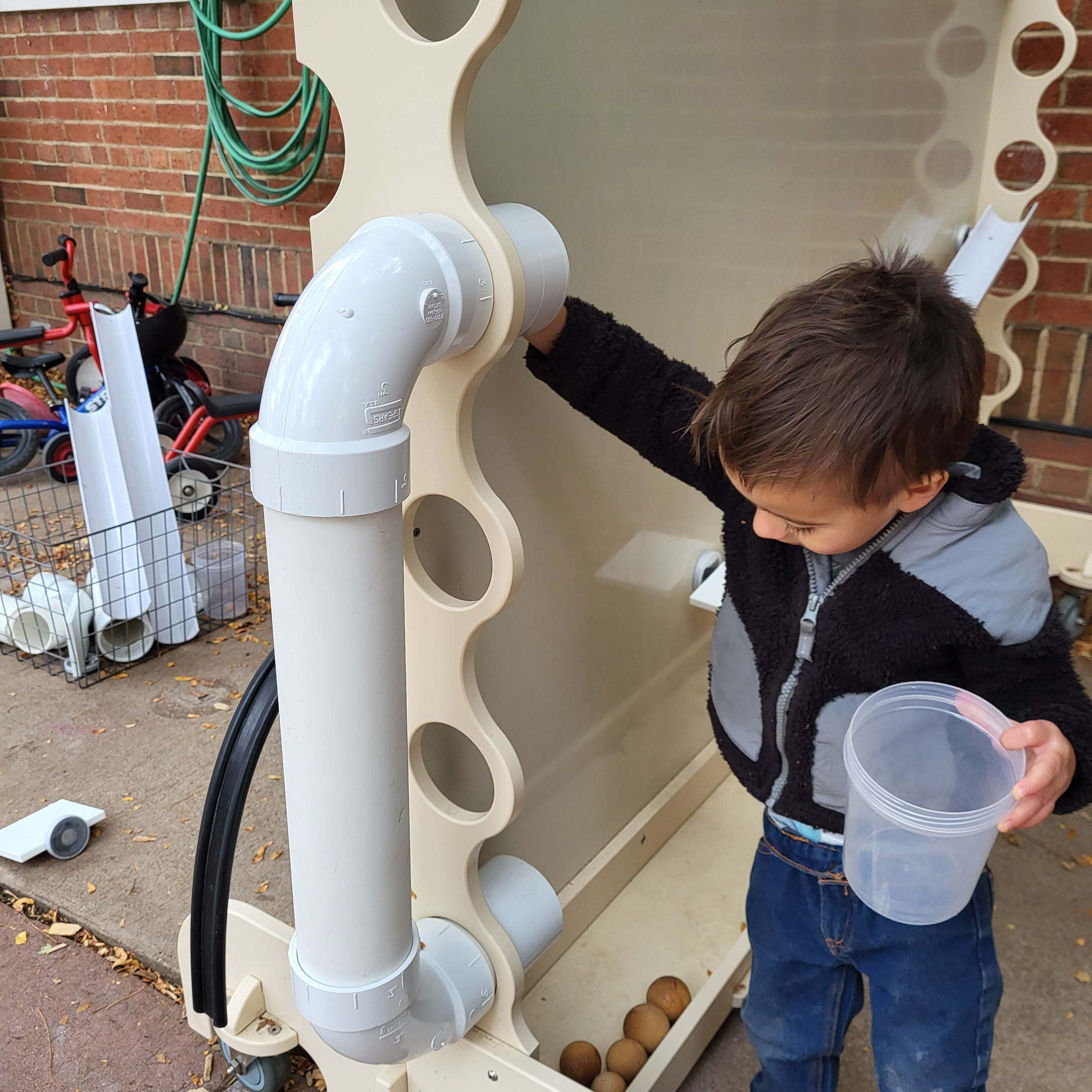 Child playing with a beige, mobile magnetic wall featuring white PVC pipes and ramps on a brick patio.