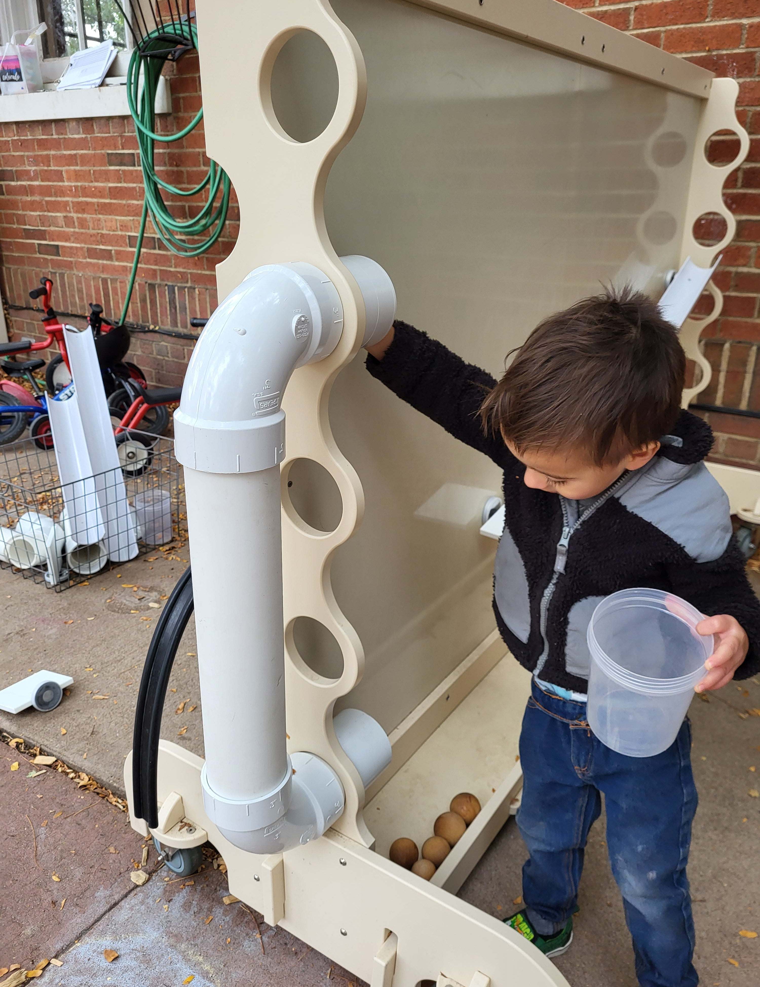 Child playing with a beige, mobile magnetic wall featuring white PVC pipes and ramps on a brick patio.