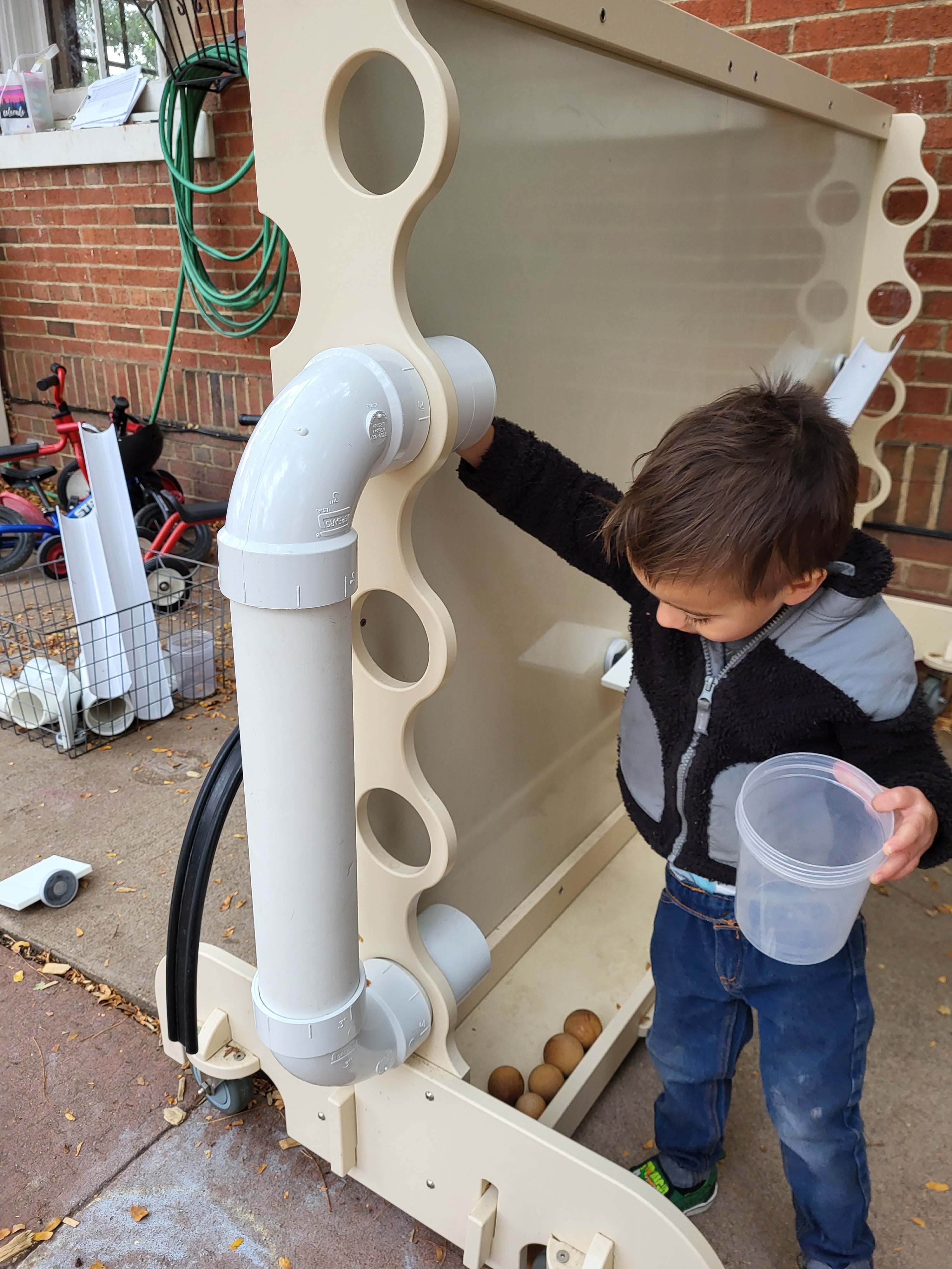 Child playing with a beige, mobile magnetic wall featuring white PVC pipes and ramps on a brick patio.