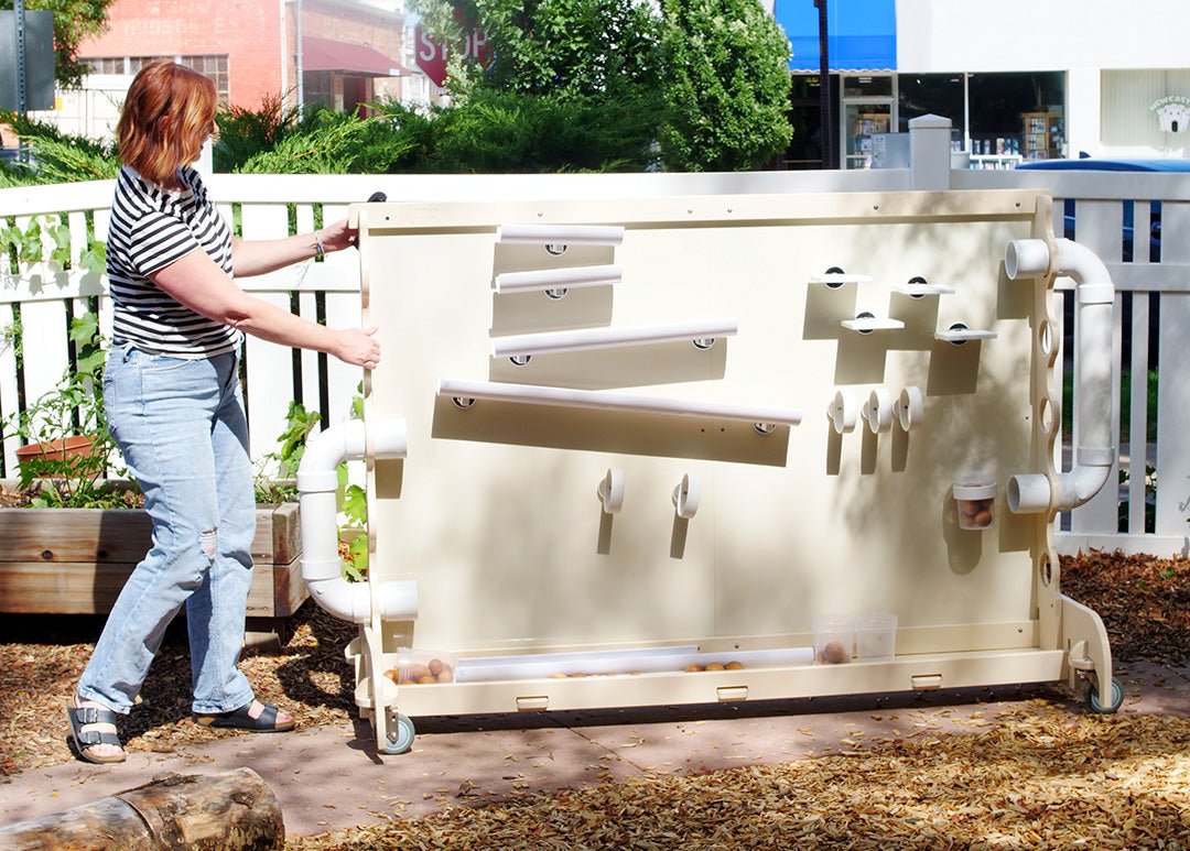 Woman moving a beige, weather-proof outdoor magnetic wall with modular ramps and tubes on a patio.