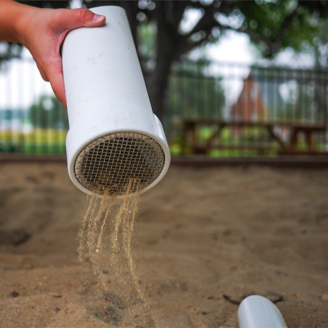 Hand holding a white cylindrical outdoor sifter scoop with a metal mesh screen, letting sand fall into a sandbox.