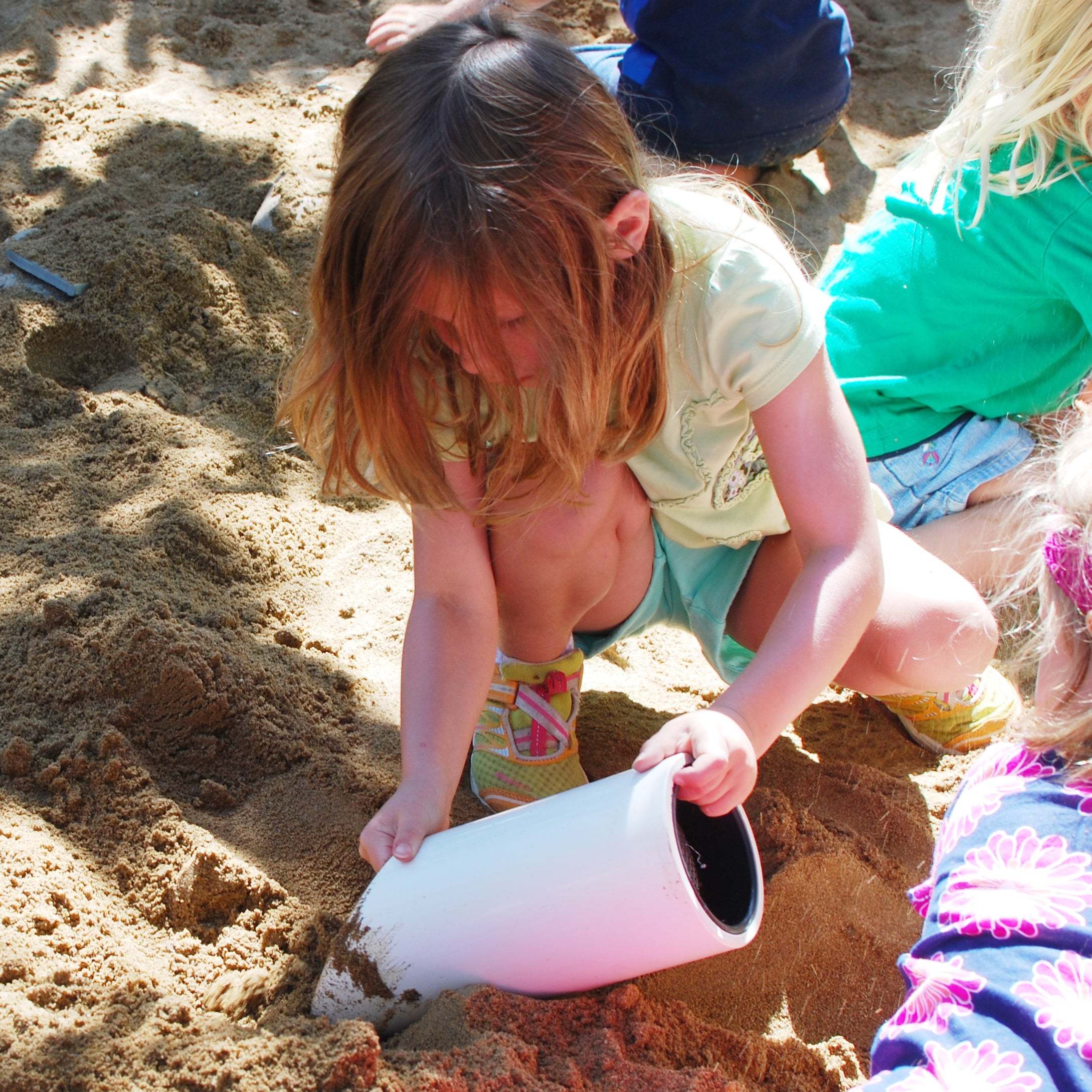 Child using a white plastic sifter scoop with a mesh screen to play in a sandbox.
