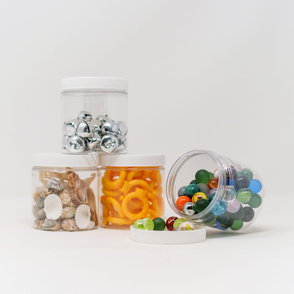 Set of four clear 8-oz plastic jars with white lids, filled with colorful sensory items like marbles, shells, and rings.
