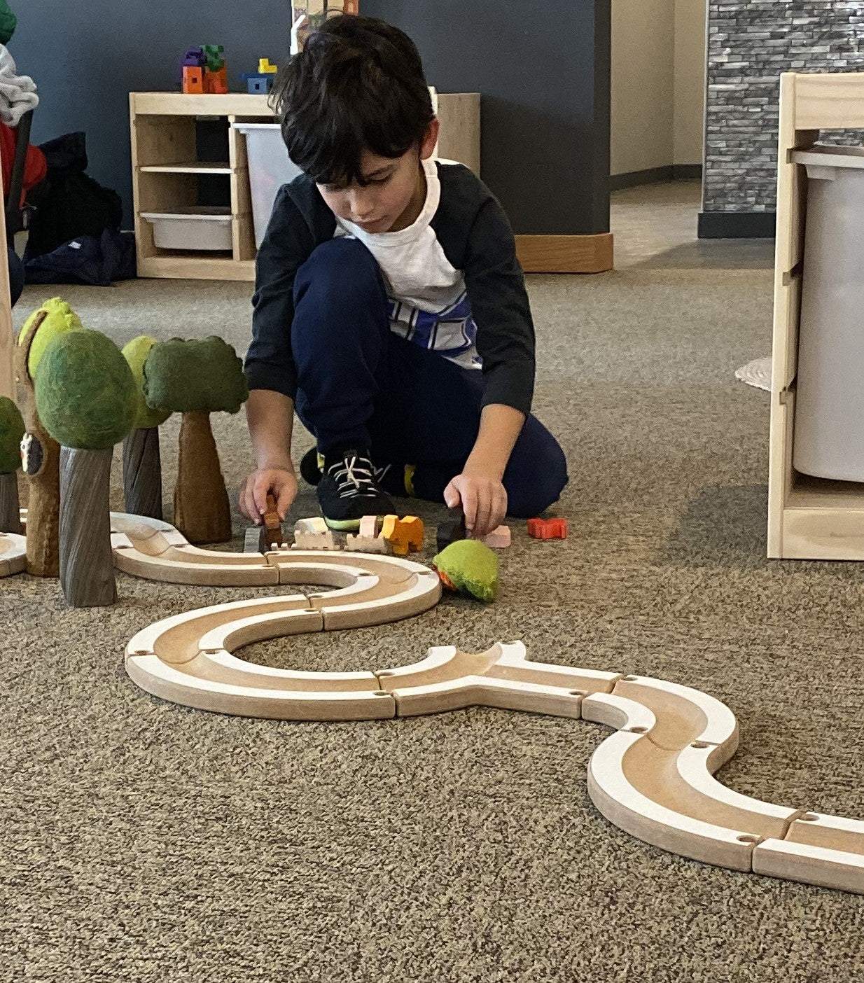 Child assembling a modular wooden puzzle ball track with felt trees and small animal figures on a carpeted floor.