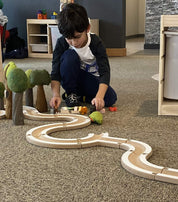 Child assembling a modular wooden puzzle ball track with felt trees and small animal figures on a carpeted floor.