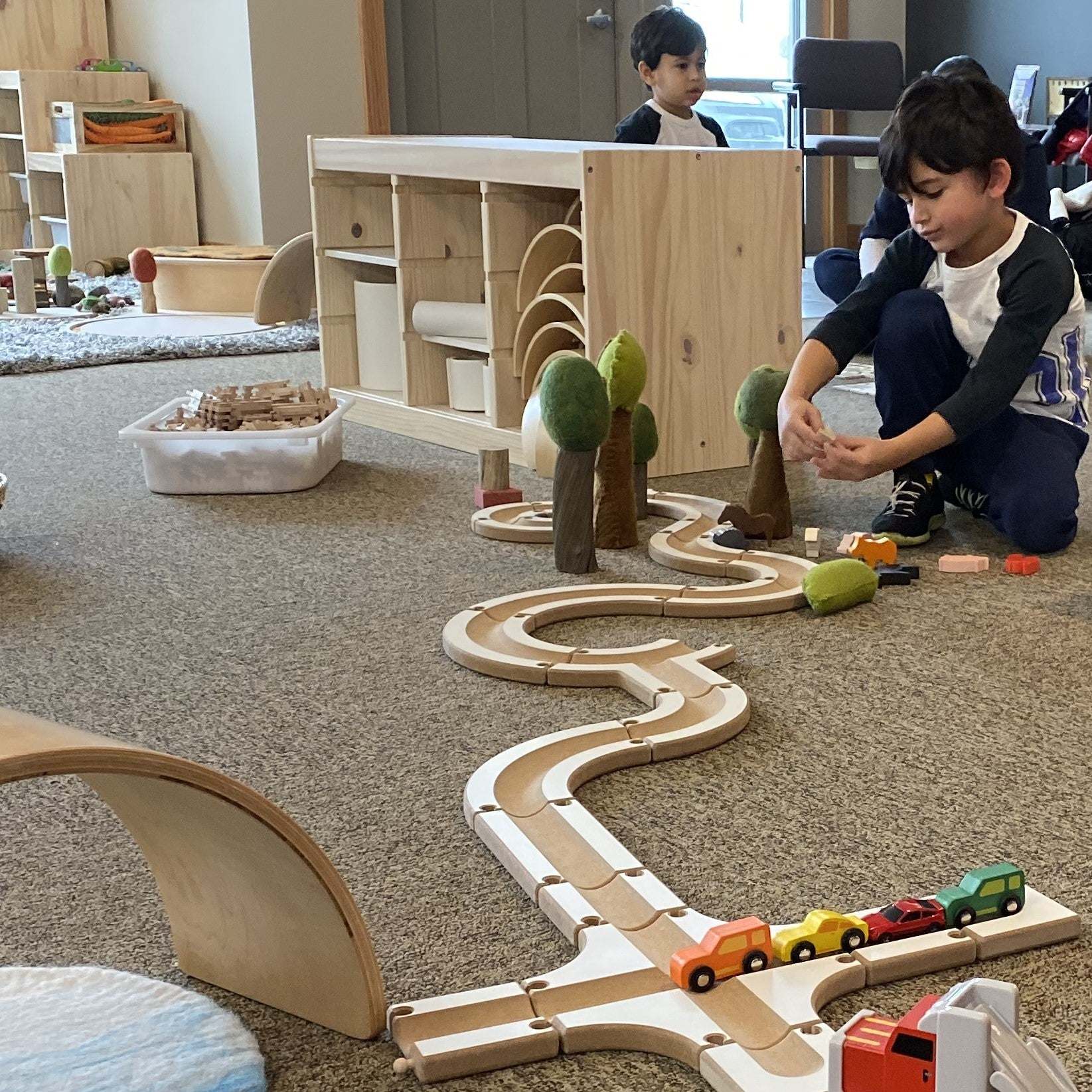 Child building a winding wooden puzzle ball track with felt trees and toy cars on a classroom carpet.