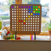 Birch wood vertical pegboard with colorful, light-catching acrylic pegs arranged on a windowsill.