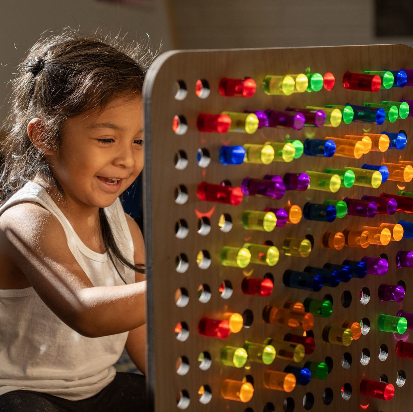 Child playing with a birch wood rainbow pegboard featuring colorful, light-catching transparent acrylic pegs.