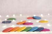 Pastel rainbow wooden discs arranged in a row on a soft white surface with twinkling fairy lights in the background.
