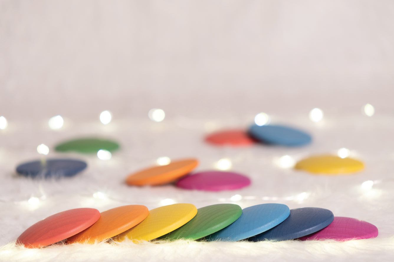 Pastel rainbow wooden discs arranged in a row on a soft white surface with twinkling fairy lights in the background.