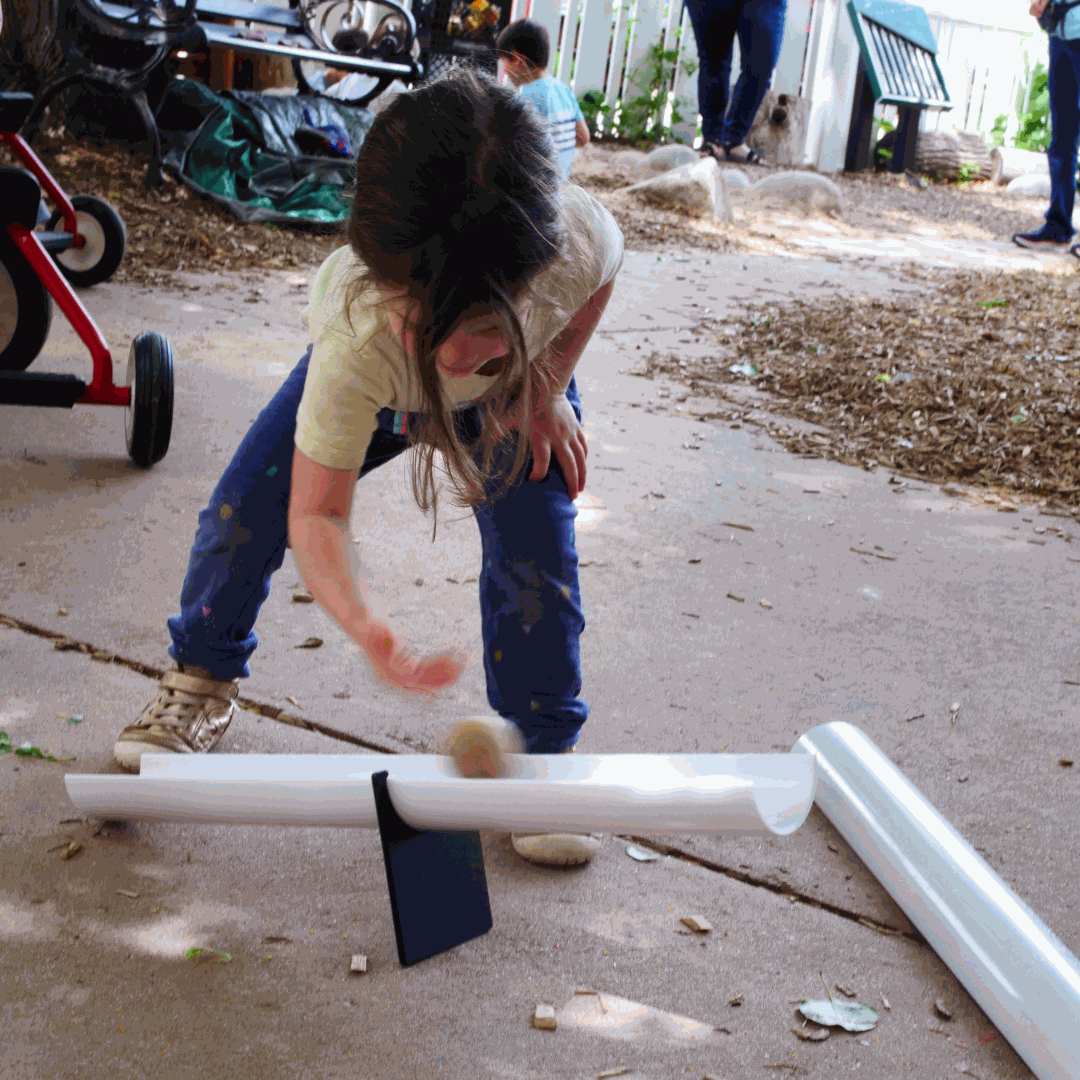 Child playing with a white plastic ramp balanced on a black metal fulcrum on a concrete patio.