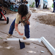 Child playing with a white plastic ramp balanced on a black metal fulcrum on a concrete patio.