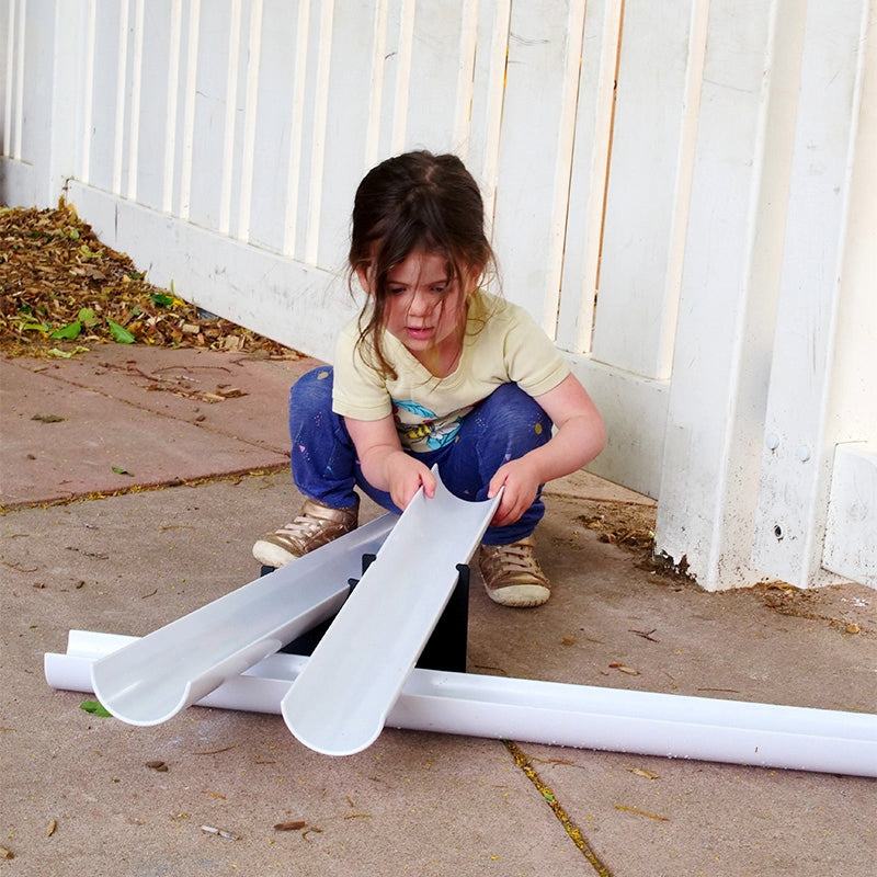 Child playing with white plastic gutter ramps and black fulcrum supports on a concrete patio.