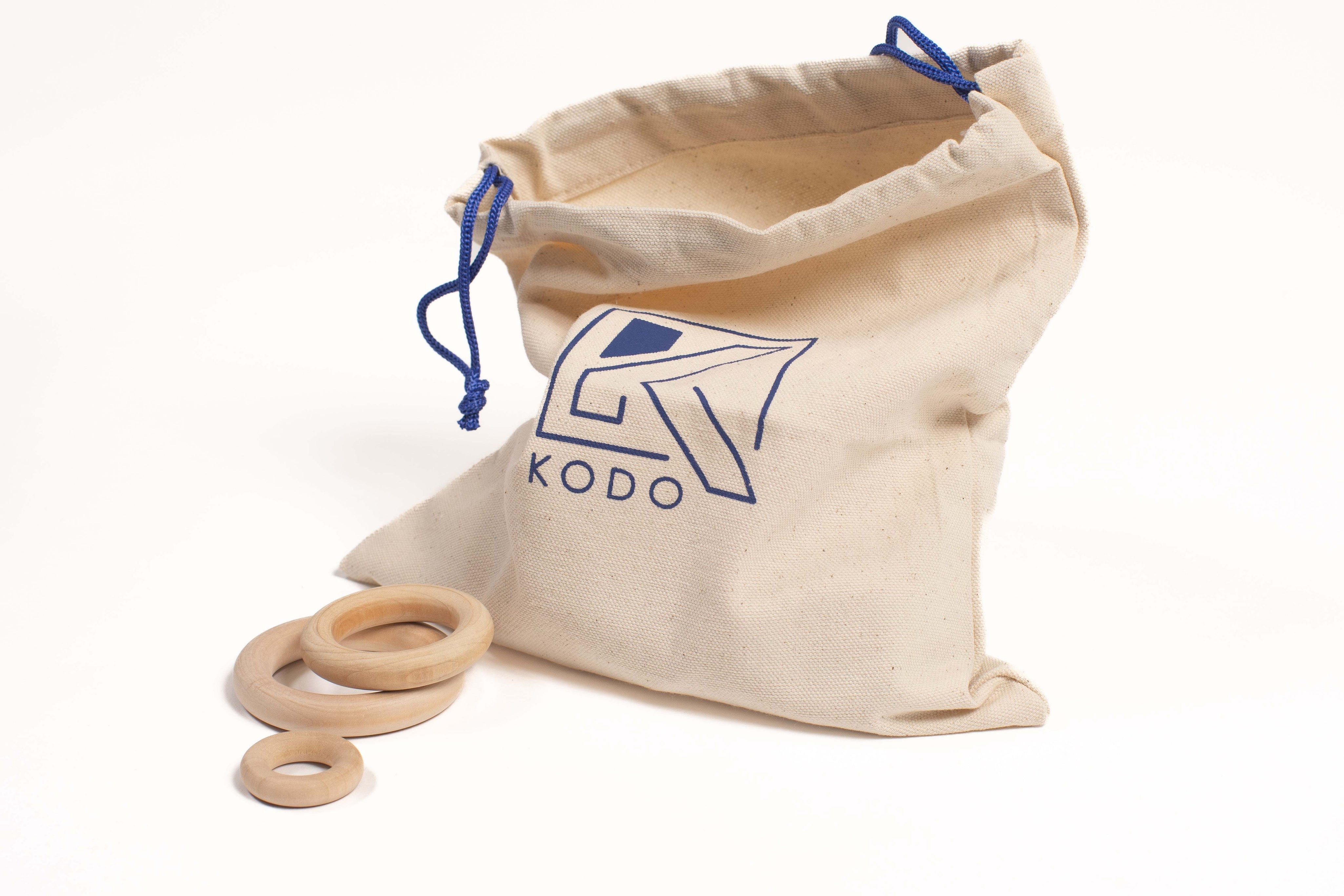 Three smooth wooden rings of varying sizes resting beside a canvas drawstring storage bag with a blue Kodo logo.