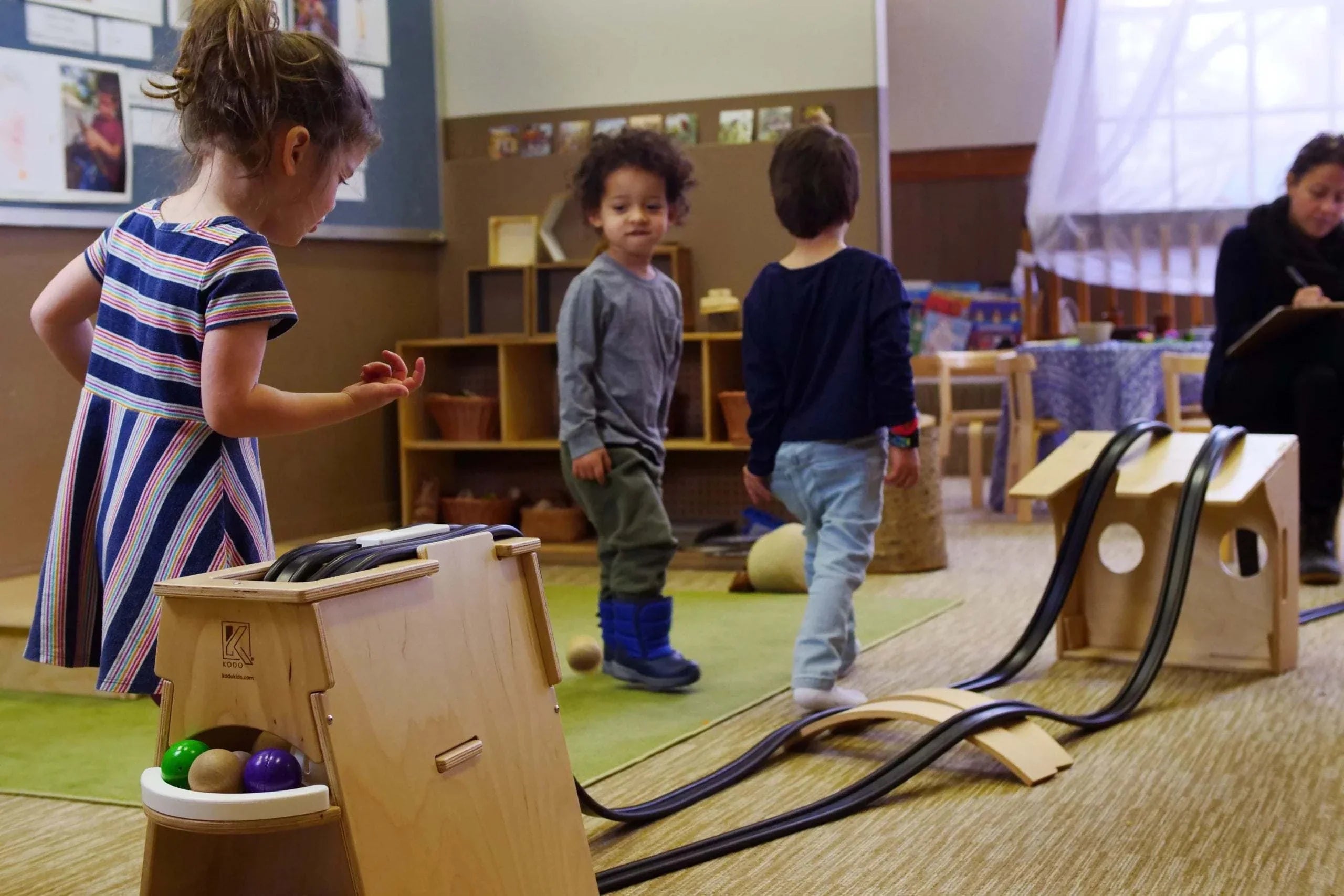 Children playing with a wooden Rubber Ramp Tower set featuring black tracks, ball storage, and elevated ramp configurations.