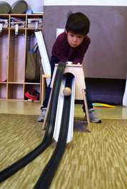 Child playing with a wooden rubber ramp tower featuring two black tracks and a ball rolling down onto a carpeted floor.